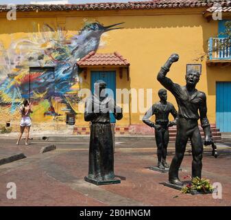 Sculture di eroi indipendenti in Plaza Trinidad di fronte all'arte di strada che decorano un edificio coloniale a Getsemani, Cartagena, Colombia Foto Stock