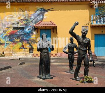 Sculture di eroi indipendenti in Plaza Trinidad di fronte all'arte di strada che decorano un edificio coloniale a Getsemani, Cartagena, Colombia Foto Stock