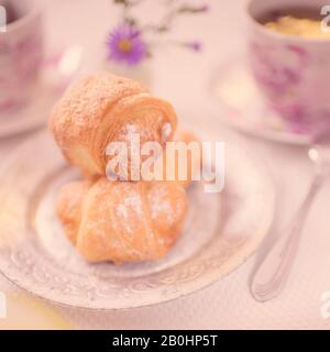 Cornetti freschi su un piatto d'argento vintage sul tavolo. Tè in tazza, cucchiaio e fiori in sfondo sfocato Foto Stock