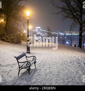 Budapest, Ungheria - panchina e lampione in un parco innevato nel quartiere di Buda con il Ponte delle catene Szechenyi sullo sfondo durante le nevicate pesanti in inverno Foto Stock