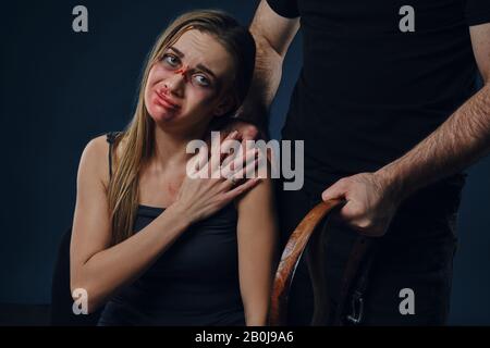 L'uomo crudele mise la mano sulla spalla femminile, mostrando la cintura. Vittima di lividi sul viso seduto nelle vicinanze. Sfondo blu. Violenza domestica. Primo piano. Foto Stock