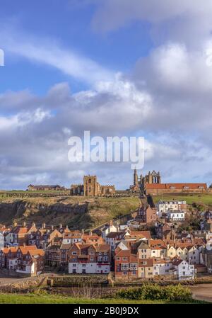 Il porto esterno di Whitby. Case annidate sotto la scogliera con una chiesa e Whitby Abbey sulla cima della collina. Un cielo nuvoloso è sopra. Foto Stock