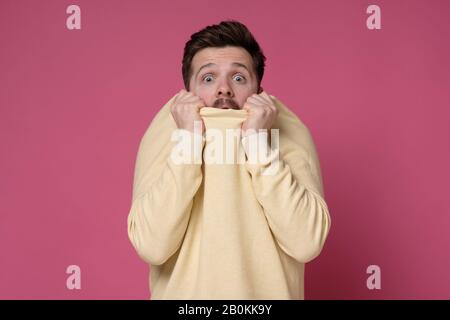 Spaventato giovane caucasico che piange attraverso il maglione. L'uomo spaventato nasconde il viso con i vestiti, guardando timido Foto Stock