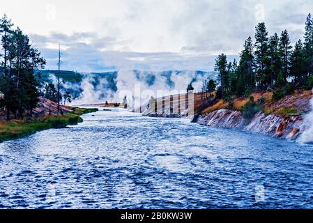 Fiume blu che scorre da geyser e colline con alberi di pino verde su entrambi i lati delle rive del fiume. Foto Stock