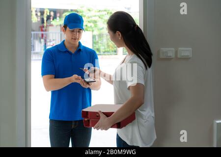 Asian consegna giovane uomo in blu uniforme sorriso e tenendo scatole della pizza in casa di fronte e donna asiatica che accetta una consegna di scatole della pizza e il pagamento Foto Stock