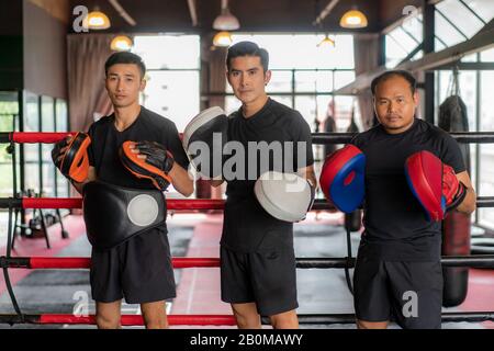 Tre pugili asiatici che guardano la macchina fotografica e posano e sorrida mentre si appoggiarono su corde rosse nere sull'anello di boxe e hanno un riposo dopo l'addestramento duro in blac Foto Stock