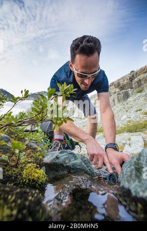 Basso angolo di uomo attivo che riempie la bottiglia d'acqua dal torrente di montagna. Foto Stock