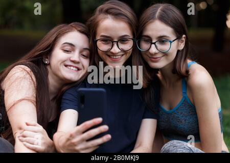 Tre ragazze sedute su tappetino yoga e facendo selfie nel parco verde Foto Stock