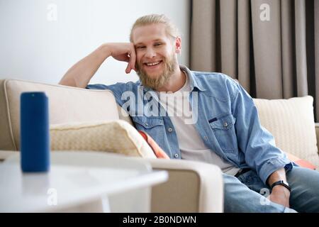 Uomo sorridente rilassarsi sul divano guardare la fotocamera utilizzare l'altoparlante wireless portatile sul tavolo Foto Stock