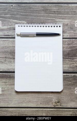 Blocco note con un foglio bianco vuoto in una carta di controllo con penna a sfera si trova sullo sfondo di tavole di legno. Foto Stock