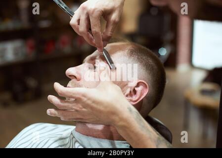 Vista ravvicinata delle mani dello stilista dei capelli rasatura della testa del cliente maschio con un rasoio da barba Foto Stock