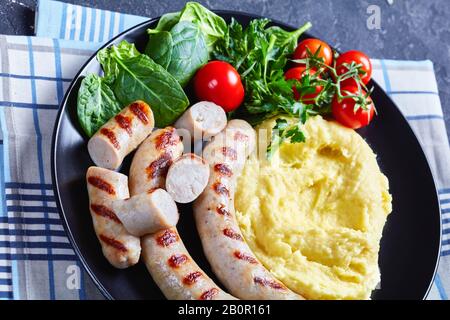 Aligot francese di patate schiacciato con formaggio fuso servito con salsicce, insalata fresca di spinaci, pomodori e foglie di insalata servita su un piatto nero con un Foto Stock