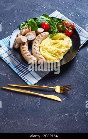 Aligot francese di patate schiacciato con formaggio fuso servito con salsicce, insalata fresca di spinaci, pomodori e foglie di insalata servita su un piatto nero, verti Foto Stock