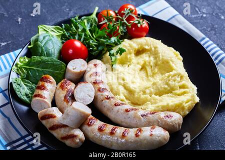 Purè di patate con scalogno e formaggio fuso servito con salsicce, insalata fresca di spinaci, pomodori e foglie di insalata servite su un piatto nero con una na Foto Stock