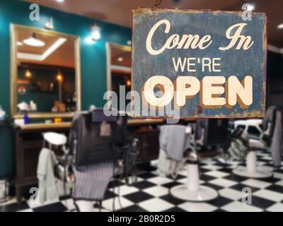 Un segno vintage di affari che dice 'come in We're Open' sulla finestra barbiere e parrucchiere negozio. Immagine di barbiere offuscata astratta e parrucchiere negozio Foto Stock