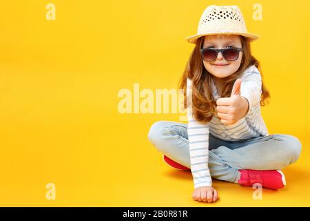 Bambina seduta sul pavimento in studio con un cappello e occhiali da sole su sfondo giallo. Il bambino mostra un pollice su. Foto Stock