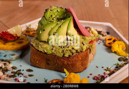 Primo piano di pane tostato con metà avocado su una base di feta e avocado strapazzate, pomodoro grattugiato e olio servito in un piatto ottagonale su tavola di legno. Artis Foto Stock