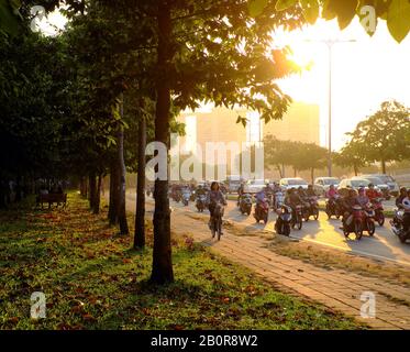 HO chi MINH CITTÀ, VIET NAM, splendida scena al mattino presto nel parco, con foglie da caduta di alberi su campo di erba, persone che si spostano sulla strada alla luce del sole Foto Stock