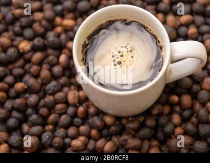 Una tazza di caffè preparato con fagioli arrostiti e macinati in modo casalingo. Espresso con panna e fagioli attorno alla tazza, spalmato su una superficie piana. Foto Stock