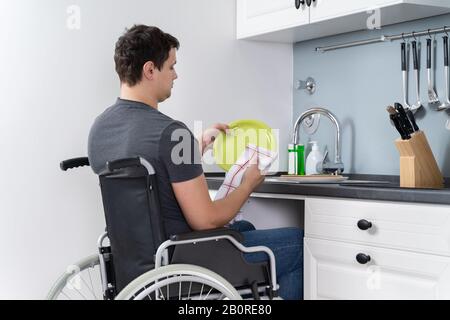 Uomo disabile seduto su una sedia a rotelle per il lavaggio e la pulizia delle stoviglie in cucina Foto Stock