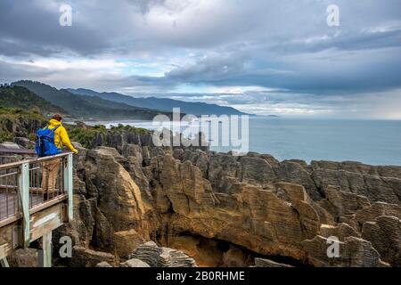 Giovane uomo sulla piattaforma di osservazione che si affaccia sul paesaggio costiero con rocce di arenaria, Pancake Rocks, Parco Nazionale di Paparoa, Punakaiki, West Coast, Sud Foto Stock
