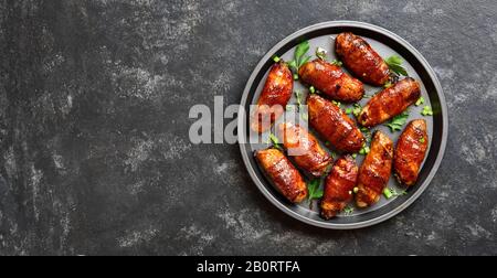 Pancetta avvolto grigliate di ali di pollo sulla piastra su pietra nera sfondo con spazio libero per il testo. Gustosi snack da carne di pollo, pancetta dolce, aspro Foto Stock