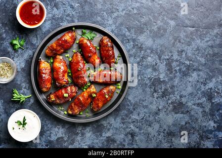 Ali di pollo alla griglia con pancetta avvolta su un piatto su sfondo di pietra blu con spazio libero. Gustoso spuntino a base di carne di pollo, pancetta in agrodolce, salate e salate Foto Stock