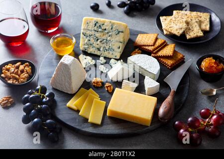 Assortimento di formaggi su tagliere in marmo scuro con vino rosso. Sfondo grigio. Foto Stock