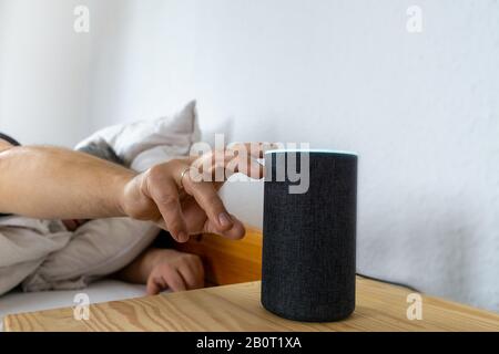 L'uomo si trova a letto e controlla il suo altoparlante intelligente Foto Stock