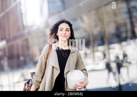 Ritratto di giovane donna con lunghi capelli marroni che indossano cappotto beige, tenendo un cappello duro, guardando la macchina fotografica. Foto Stock