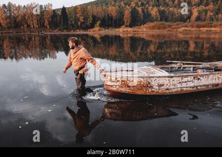 Uomo che trascina vecchia barca arrugginita in mezzo al lago in autunno, Russia Foto Stock