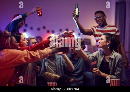Gruppo di amici felici che tengono tazze e brindano con la birra mentre celebrano insieme a casa festa Foto Stock