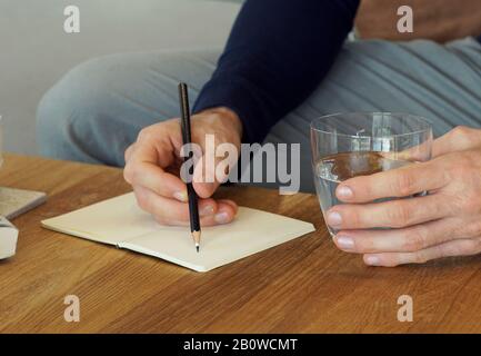 Uomo seduto su un divano che scrive in un notebook e tiene un bicchiere d'acqua Foto Stock
