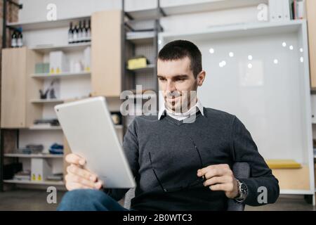 Uomo felice seduto e tenuto un tablet. Sorridente lavoratore di ufficio che riposa e tiene i suoi occhiali mentre guarda il suo tablet nello spazio di coworking. Foto Stock