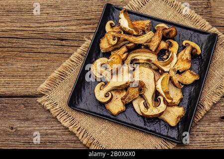 Funghi porcini secchi. Ingrediente tradizionale per cucinare cibi sani. Piastra in ceramica nera, tovagliolo in tessuto, vecchi pannelli in legno sfondo, copia sp Foto Stock