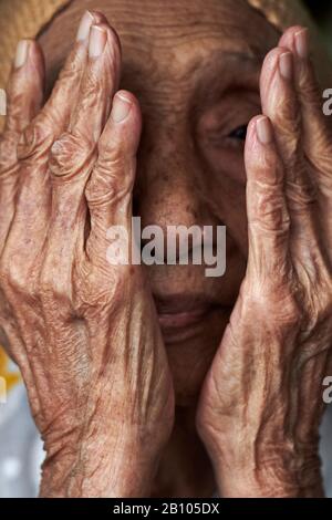 Ritratto di una donna malese anziana che copre il suo viso con le sue mani rugose Foto Stock