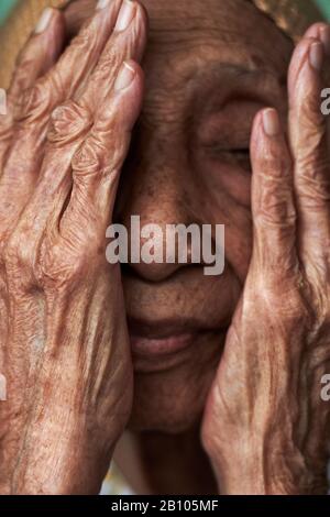 Ritratto di una donna malese anziana che copre il suo viso con le sue mani rugose Foto Stock