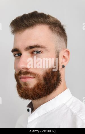 Immagine closeup di giovane bell'uomo con la barba posa e guardando la fotocamera isolato su sfondo grigio Foto Stock