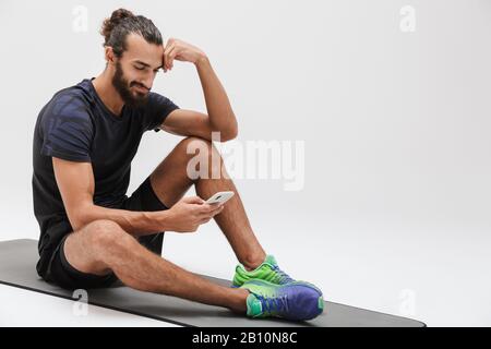 Immagine di sportivo bruna atletico in un tracksuit che tiene lo smartphone mentre si siede su tappetino yoga isolato su sfondo bianco Foto Stock