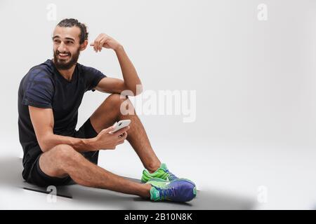 Immagine di Happy brunette sportivo in tuta tenendo lo smartphone mentre si siede su tappetino yoga isolato su sfondo bianco Foto Stock