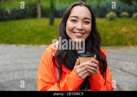 Giovane donna asiatica sorridente che indossa un impermeabile camminando all'aperto sotto la pioggia, tenendo una tazza di caffè Foto Stock