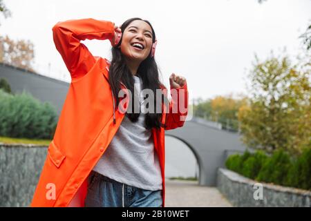 Giovane donna asiatica sorridente che indossa un impermeabile che cammina all'aperto sotto la pioggia, ascoltando musica con cuffie wireless Foto Stock