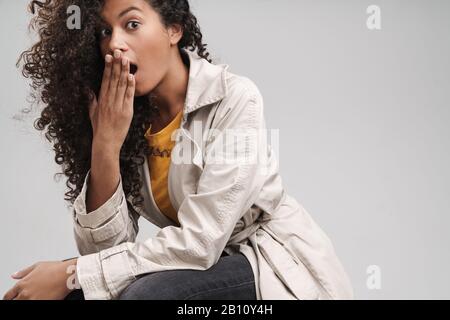 Primo piano di un attraente scioccato giovane donna africana con capelli ricci, indossando cappotto autunno isolato su sfondo grigio Foto Stock