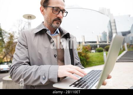 Immagine di un uomo d'affari concentrato con bearded in occhiali usando un laptop mentre si sta in piedi in strada della città Foto Stock