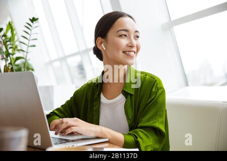 Sorridente bella giovane donna asiatica che lavora sul computer portatile mentre si siede al caffè all'interno, guardando attraverso i documenti Foto Stock