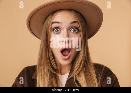 Immagine di giovane sorpresa ragazza indossare cappello e cappotto che esprime meraviglia con bocca aperta isolato su sfondo beige Foto Stock