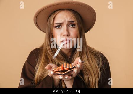 Immagine di una ragazza caucasica malata che indossa cappello che tiene il mazzo di pillole con termometro nella sua bocca isolato su sfondo beige Foto Stock