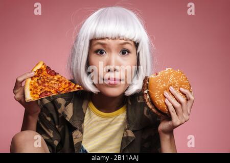 Immagine di giovane ragazza asiatica in bianco parrucca scelta mentre tenendo fetta di pizza e hamburger grande isolato su sfondo rosa Foto Stock