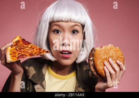 Immagine di giovane ragazza asiatica in bianco parrucca esitante mentre tiene fetta di pizza e grande hamburger isolato su sfondo rosa Foto Stock