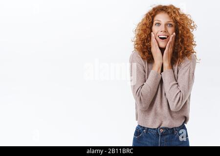 Affascinata, stupita redhead curly caucasica donna sospira da fascino e gioia, toccante guancia, sguardo si chiedeva e divertito vedere qualcosa di stupefacente Foto Stock